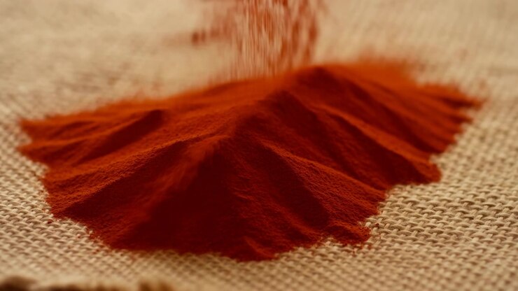 Close view shows reddish powder forming soft ridges on woven textured background while fine particles change shape gradually under warm lighting with minimal abstract arrangement