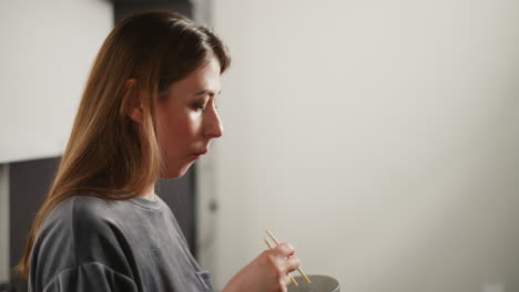 side view of woman in casual gray shirt eating noodles with chopsticks, standing in bright modern kitchen, soft blur background with white walls and cabinets
