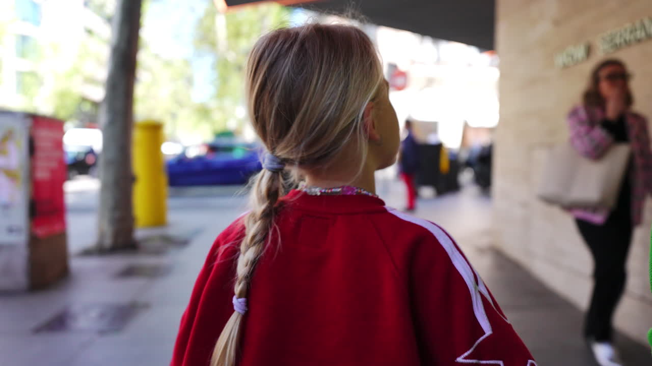 Blond haired girl wearing red sweater walking alongside parent, holding hands on sunlit Madrid street, capturing intimate family moment of urban exploration and childhood connection