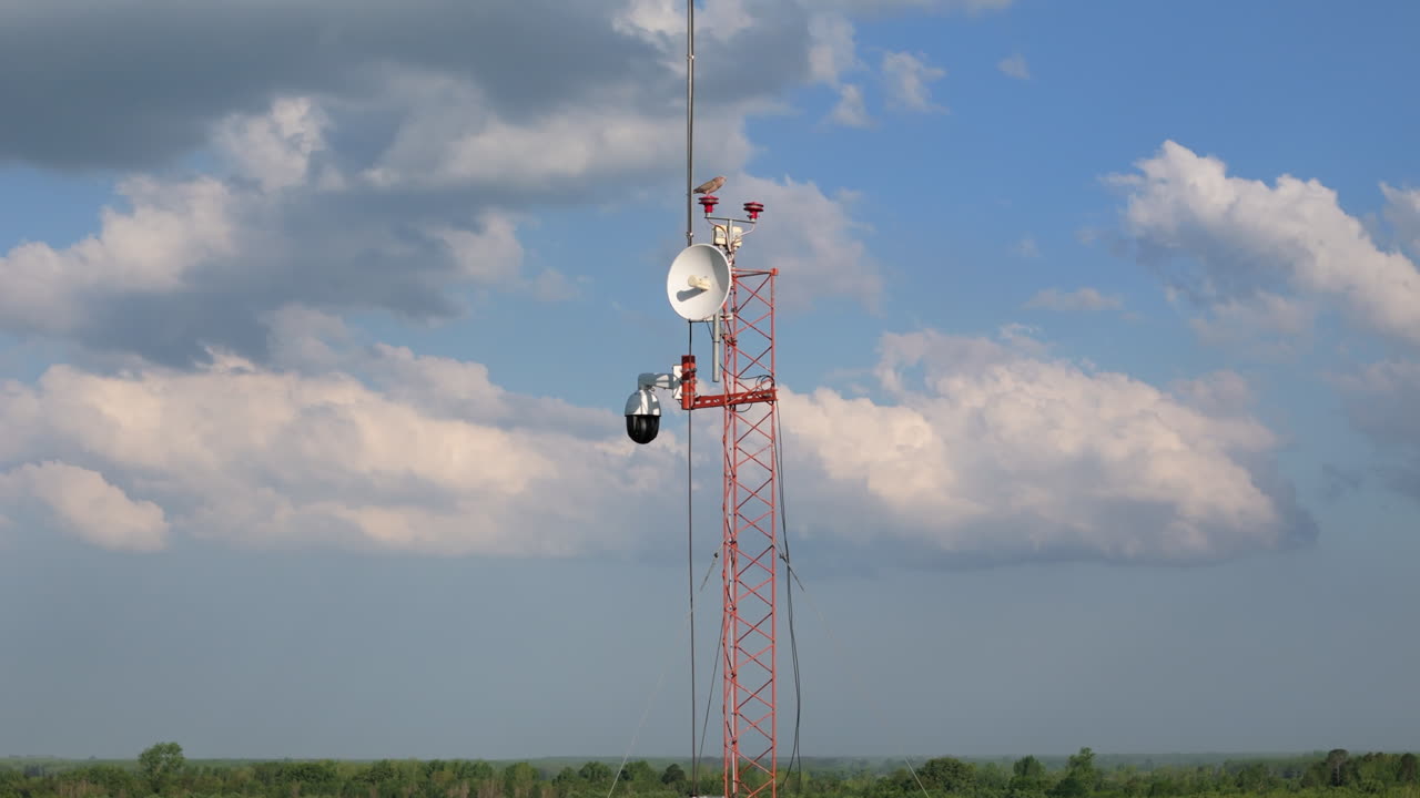 Drone footage capturing telecommunications tower with satellite dishes and antennas. Close-up aerial video of cellular network infrastructure against backdrop of white fluffy clouds.