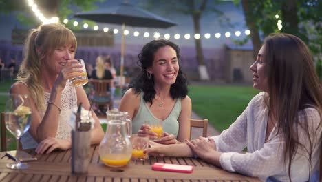 Three-Elegant-Female-Friends-Sit-In-The-Outdoors-Cafe,-Drink-Juice-And-Have-Fun-Communicating