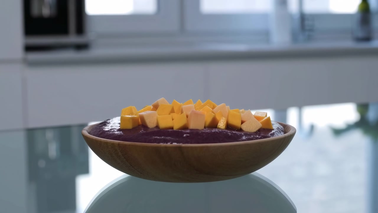 Una piccola ciotola di legno piena di cubetti arancioni rimane su un tavolo di vetro vicino alla finestra, evidenziando riflessi lisci sulla superficie, luce naturale, una disposizione semplice e un ambiente interno pulito con messa a fuoco morbida.