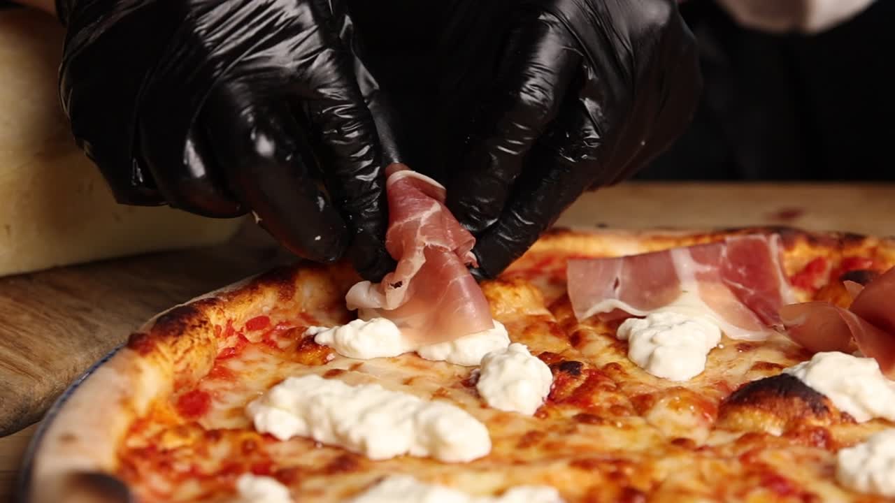 Chef con guanti neri che adagia prosciutto e ricotta su una pizza appena sfornata