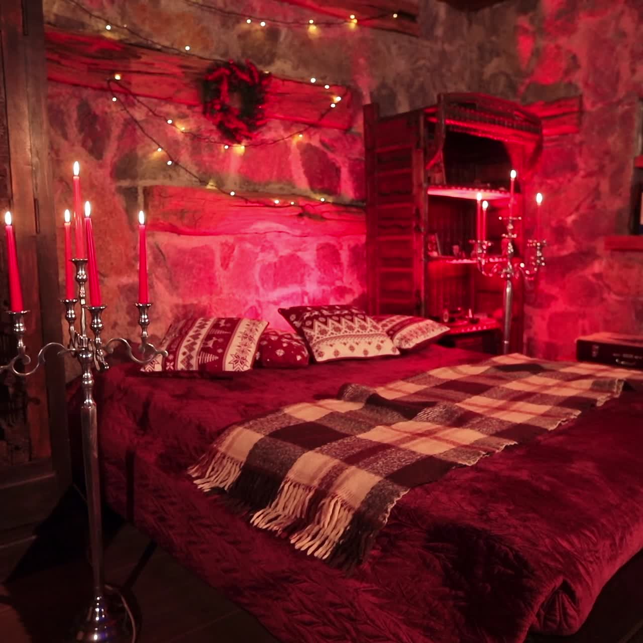 Big double bed in the beautifully decorated room with red lights. Stone walls, hard wooden old furniture and candles lighted in the countryside house bedroom