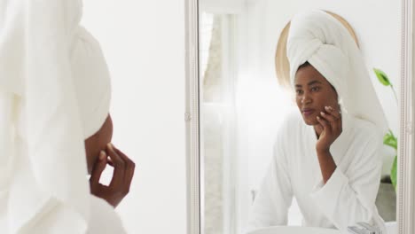 Video-Einer-Glücklichen-Afroamerikanischen-Frau-In-Robe,-Die-In-Den-Spiegel-Blickt-Und-Ihr-Gesicht-Mit-Feuchtigkeit-Versorgt