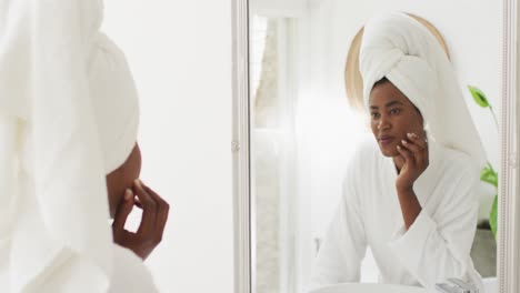 video de una feliz mujer afroamericana en túnica mirando al espejo y hidratando la cara