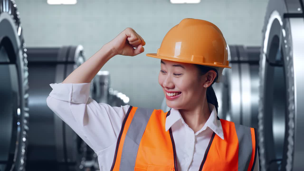 안전 헬을 착용한 아시아 여성 엔지니어가 금속 공장에서 서서 팔다리를 구부리고 카메라에 미소 짓고 있습니다.