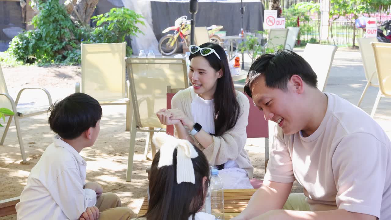 ภาพระยะกลางแบบถือถ่ายของพ่อแม่ที่ยิ้มแย้มปรบมือและพูดคุยกับเด็กสองคนในร้านกาแฟกลางแจ้งที่มีแสงแดดส่อง