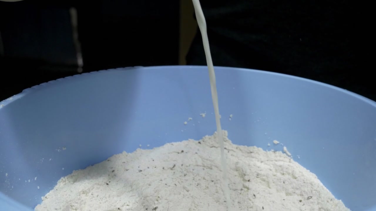 Susu dituangkan dari teko ke dalam mangkuk biru berisi tepung terigu dan biji adas manis. Latar belakang gelap. Kamera mengikuti aliran susu.