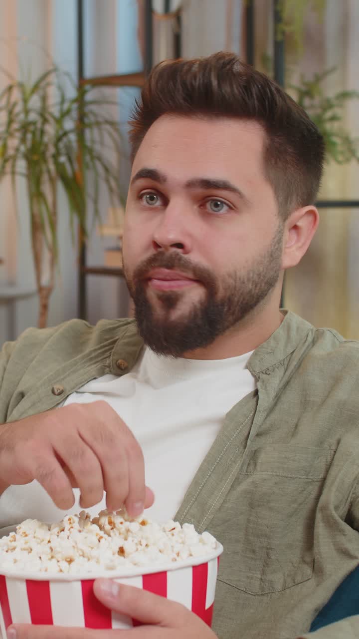 hombre moreno guapo comiendo palomitas de maíz y viendo películas sentado en el sofá en el apartamento de casa