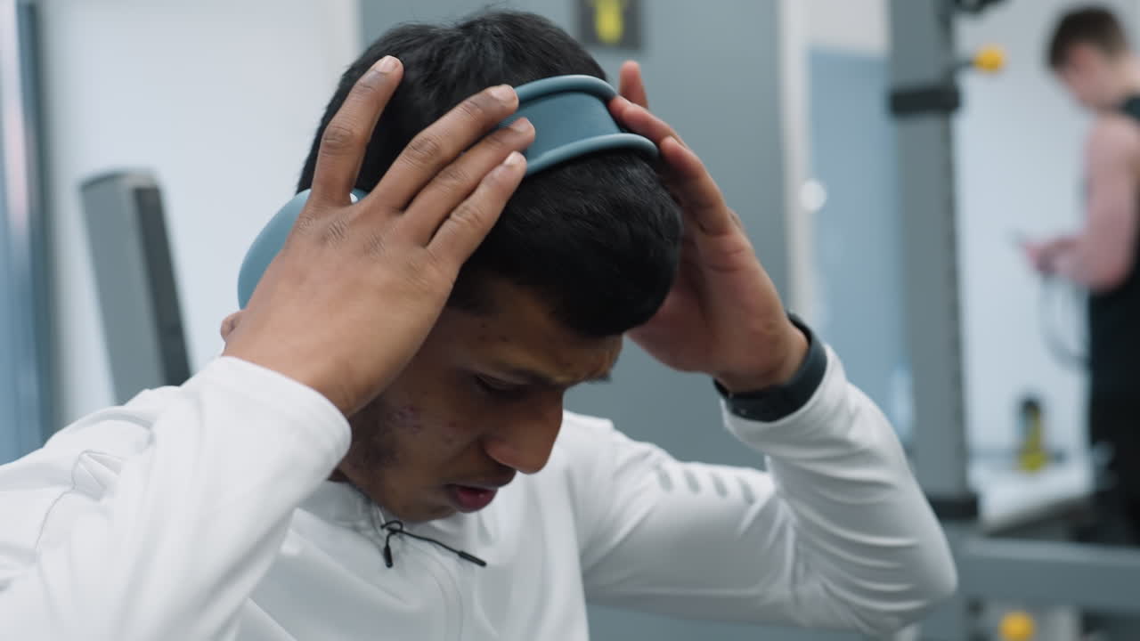 Close up of gym rat adjusting headphone while lying on bench under barbell before lift window behind shows car passing outside gym floor setup focus anticipation simple workout routine