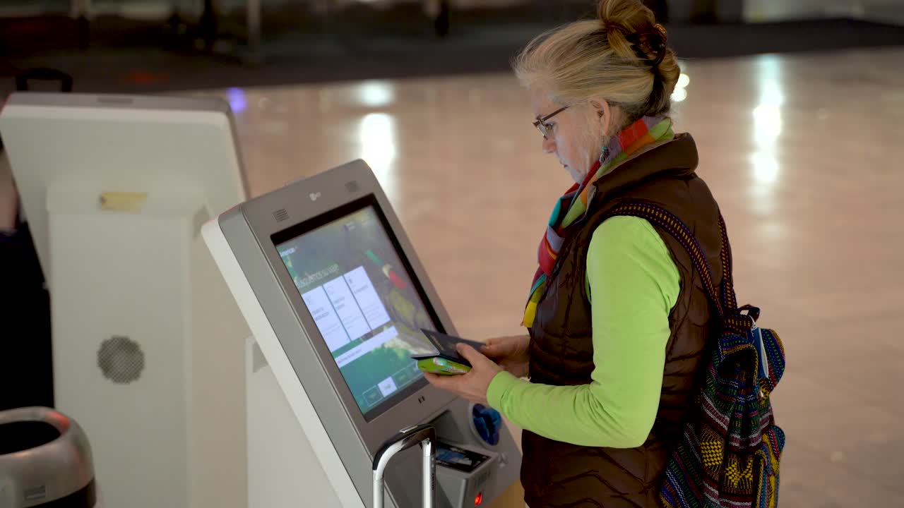 With passport and smartphone in hand, and mature woman walks up to an airport self check in terminal and checks herself in