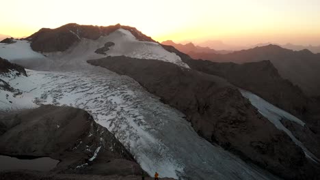 aerial flyover over two hikers enjoying the view on a cliff above glacier de vouasson near arolla in valais, switzerland at sunset with colorful glow behind layers of mountains