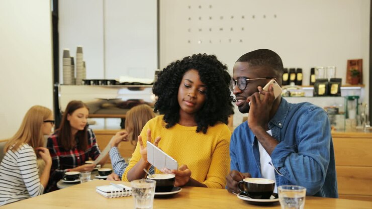 homme afro-américain parlant au téléphone et femme afro-américaine regardant une vidéo sur une tablette assis à une table dans un café