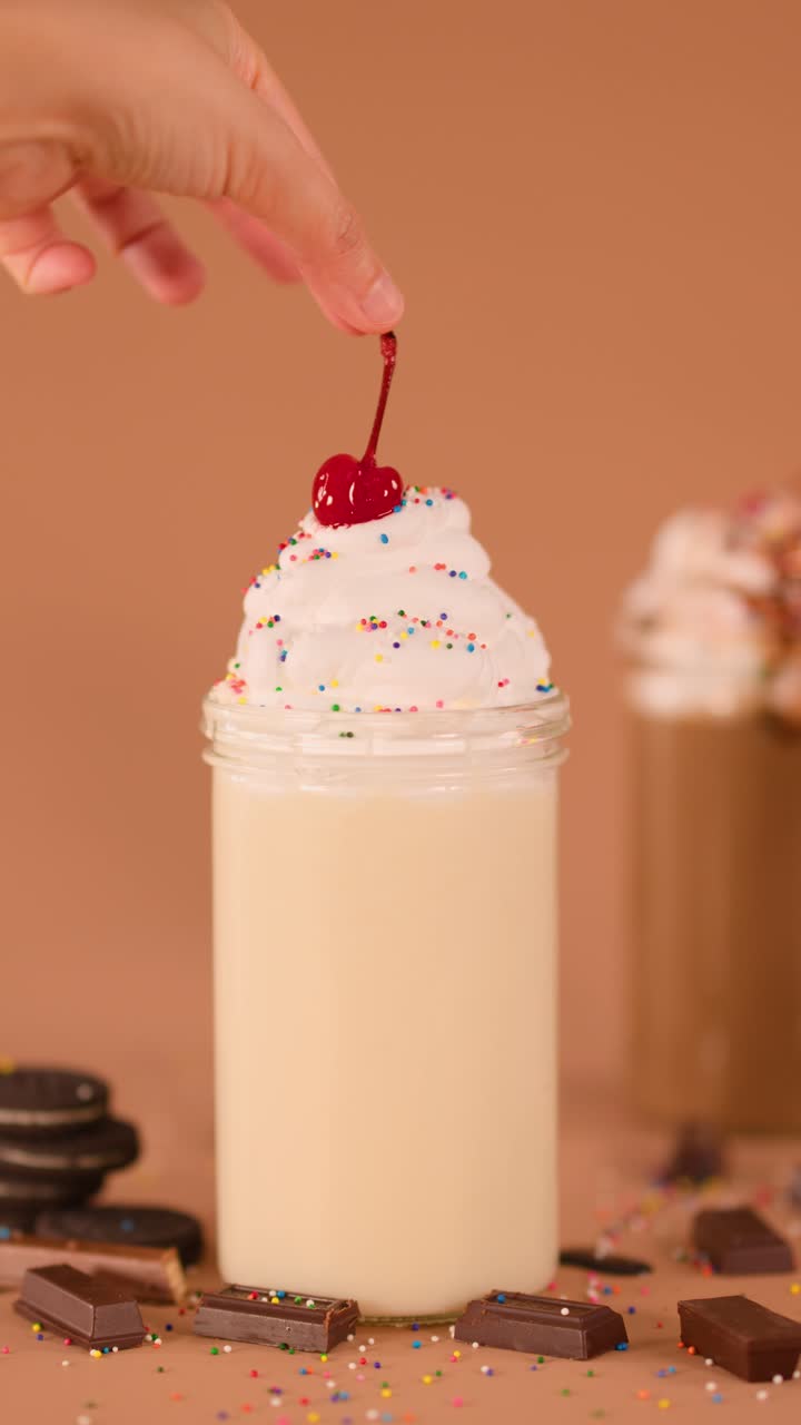 Primo piano di una mano che guarnisce un cremoso milkshake con ciliegia, zuccherini colorati, biscotti e cioccolato.
