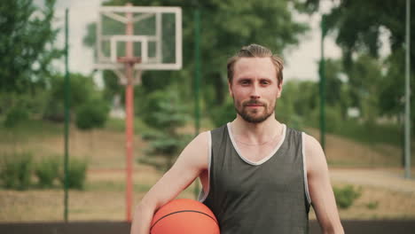 retrato de un apuesto jugador de baloncesto rubio barbudo sosteniendo una pelota, mientras mira la cámara y sonríe con confianza en una cancha de baloncesto al aire libre