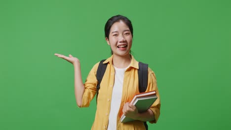 asian woman student with a backpack and some books smiling and pointing to side while standing in the green screen background studio