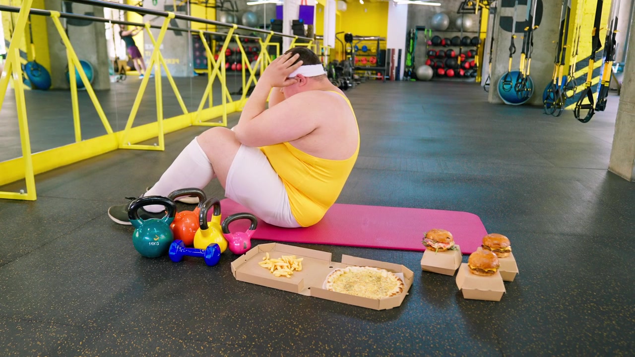 Uomo in sovrappeso che fatica a fare addominali accanto a del fast food in palestra