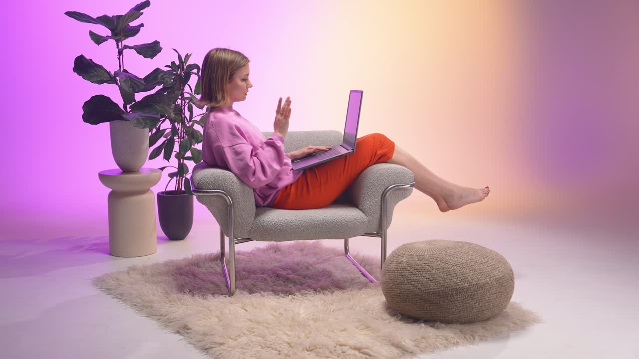 Young woman sits barefoot in a cozy armchair, working on a laptop. Surrounded by modern decor, plants, and soft pastel lights, she enjoys a relaxed and focused remote work environment