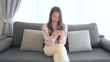 happy woman typing on a smartphone with two hands asian adult in casual clothes sitting on the sofa and typing with both hands message in mobile phone