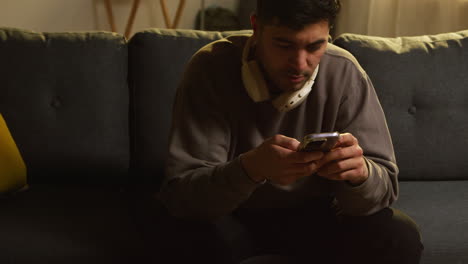 joven con auriculares inalámbricos pasando la noche en casa sentado en el sofá con el teléfono móvil desplazándose por internet o redes sociales