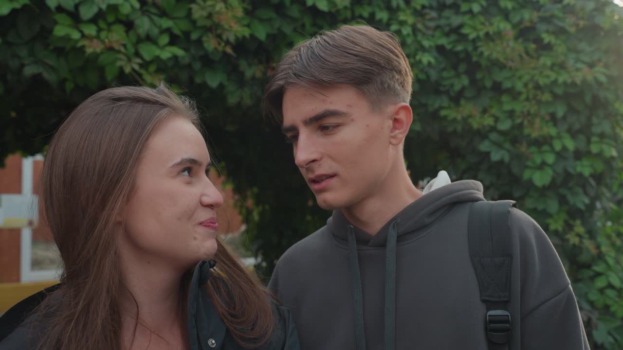 Adolescenti bianchi che si scambiano sguardi giocosi, primi piani con felpa e zaino, flirt sottile e risate leggere, contatto visivo dinamico, reazioni espressive, sfondo ricoperto di edera, spontaneo