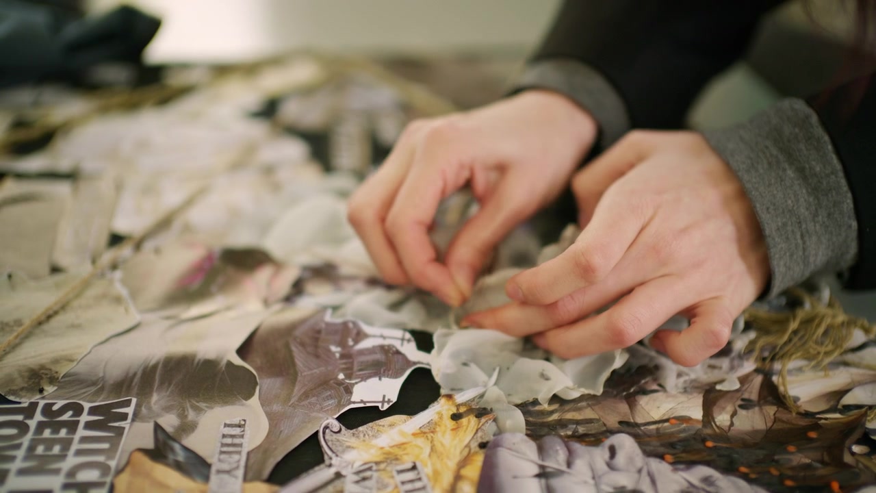 Primo piano di una mano che sistema materiali per una mood board di moda su un tavolo, inclusi bozzetti di capi stampati, campioni di tessuto e pezzi di plastica testurizzati.