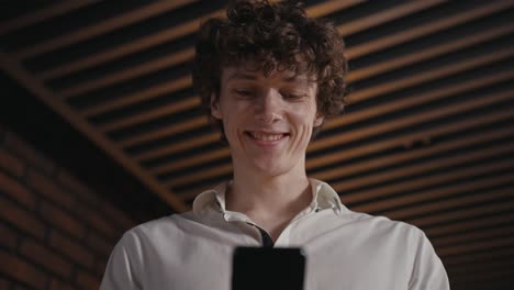 Close-up-of-a-happy-guy-with-curly-hair-in-a-white-shirt-looking-at-the-screen-of-his-smartphone-and-smiling-widely-in-the-office