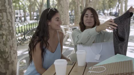 mujer latina mostrando camisa a un amigo de pelo largo en la terraza de una cafetería