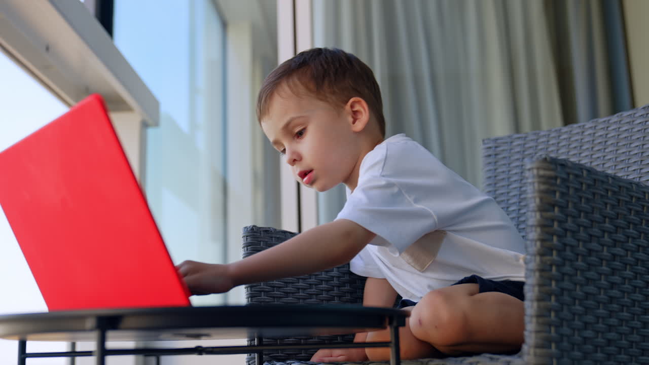 Child engaged with laptop on balcony. A young boy sits on a balcony, focused on his laptop, enjoying a quiet moment in the sun