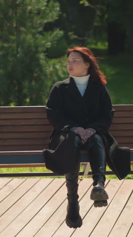 joven con abrigo negro se sienta serenamente en un columpio de madera en un parque iluminado por el sol, manos entre las piernas, mirando a la distancia, rodeada de vegetación