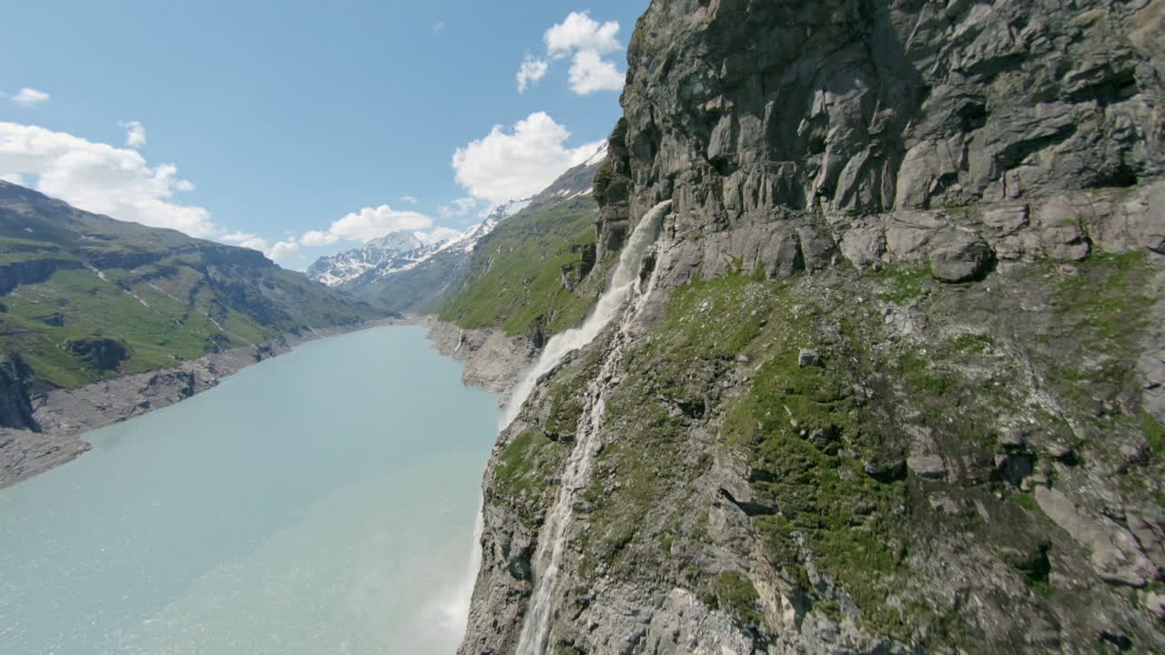 Stunning FPV and drone shots soaring through the Swiss Alps, capturing the Mauvoisin Dam and its majestic waterfall on a clear, sunny summer day.