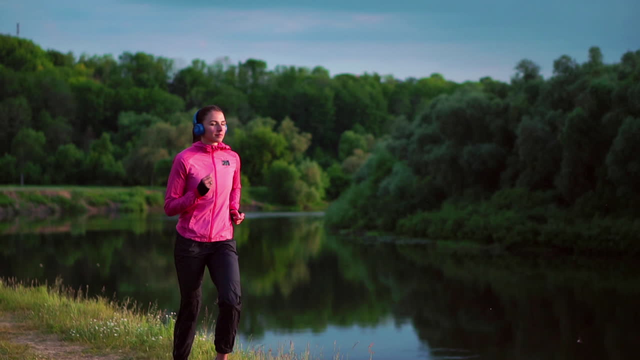 A girl in a pink jacket and black pants runs near the river in headphones preparing for the marathon