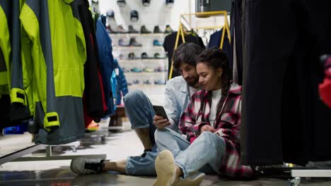 Couple-in-the-sport-department-store-sitting-on-the-floor,-watching-mobile-phone