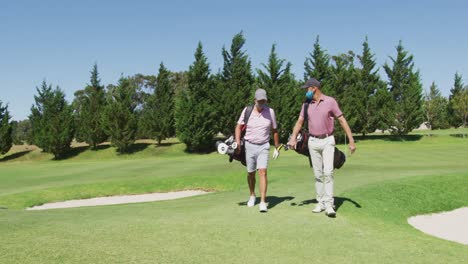 Two-caucasian-senior-man-wearing-face-masks-walking-with-their-golf-bags-at-golf-course