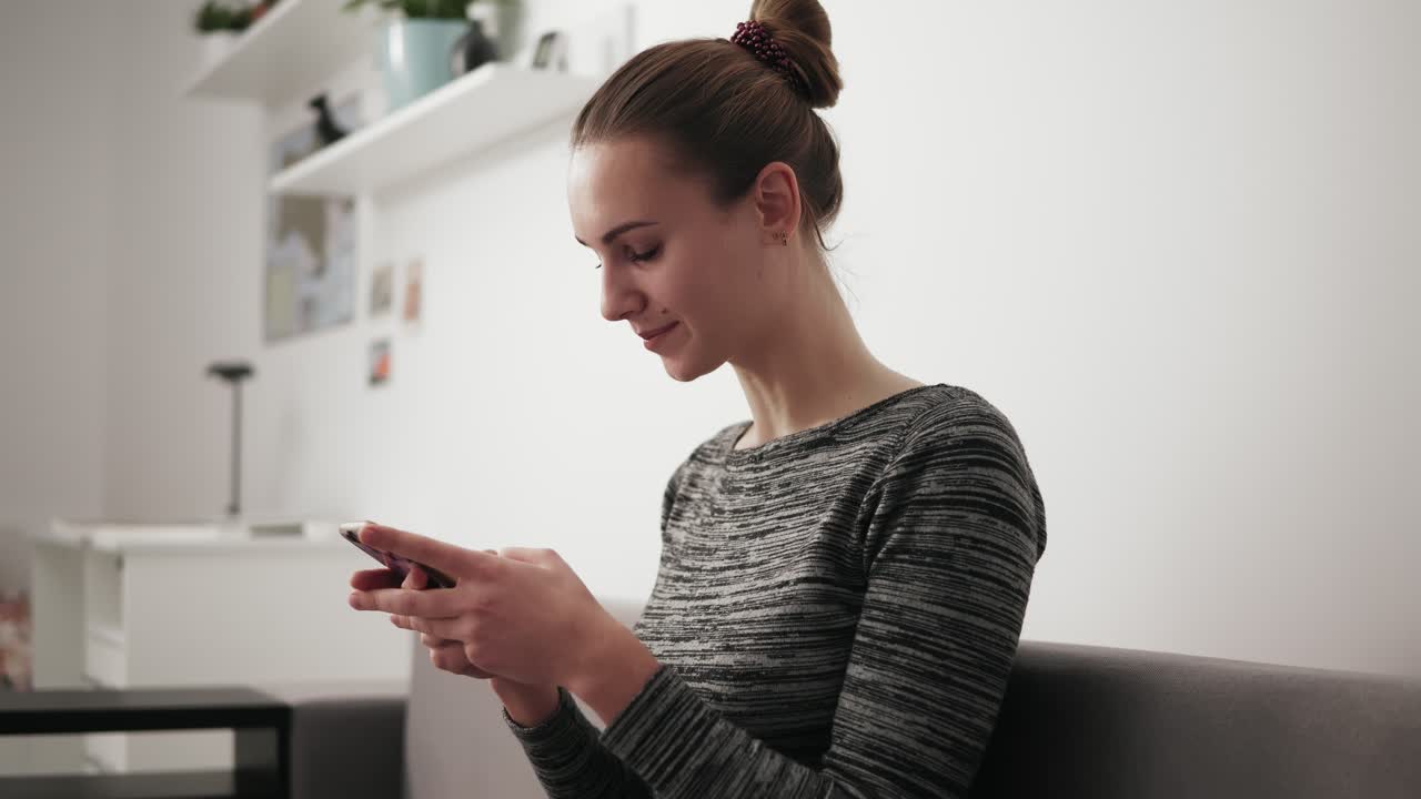 hermosa mujer joven sentada en el sofá en casa mirando el teléfono con una sonrisa y escribiendo un mensaje