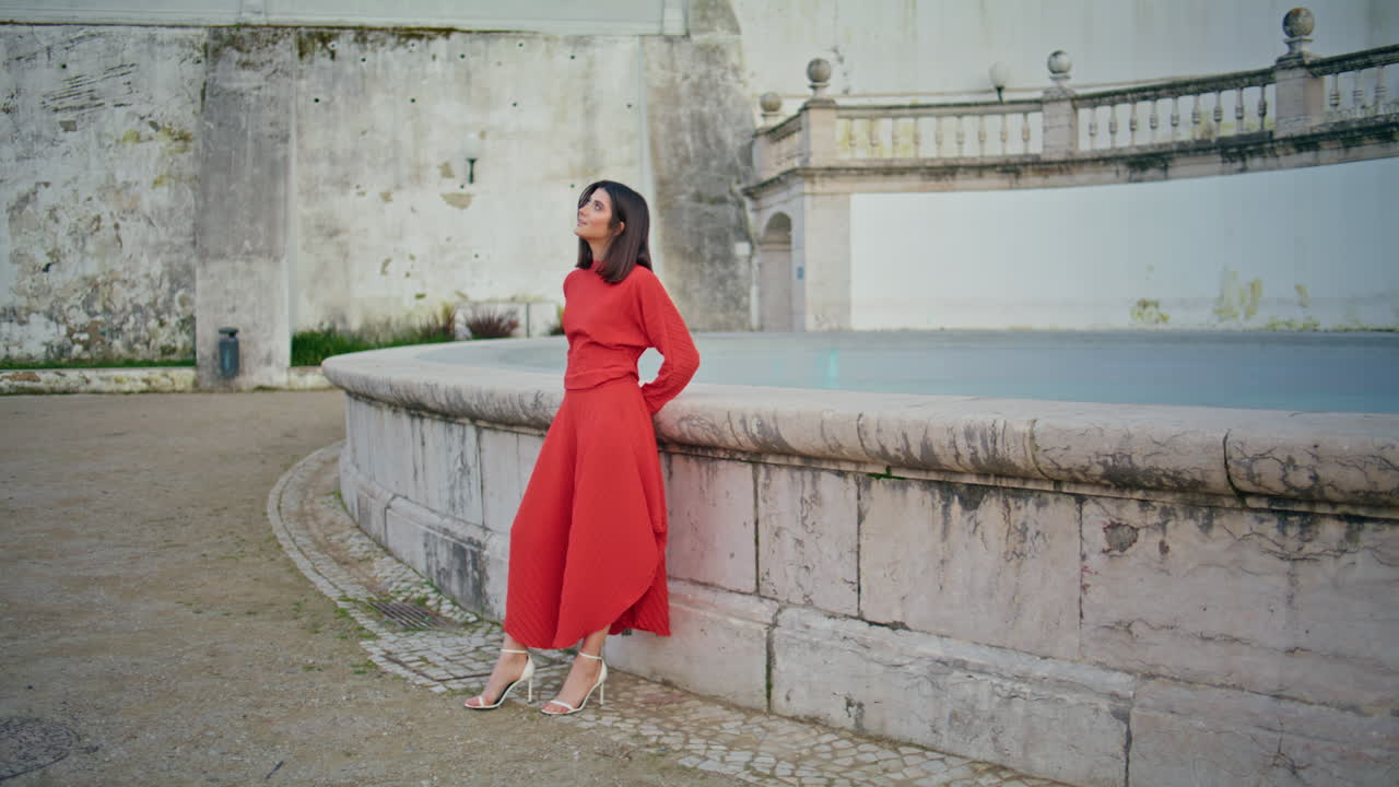 Gorgeous woman posing fountain at city looking distance. Model in red dress