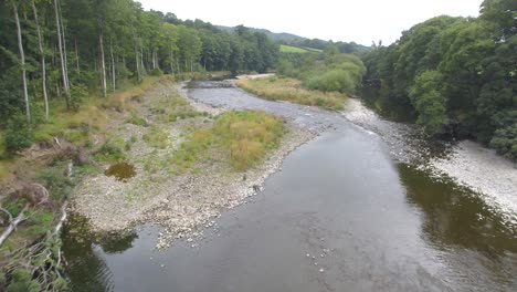 Beautiful-drone-footage-flying-slowly-and-low-between-and-over-a-tree-lined-shallow-slow-moving-meandering-river-and-river-island-in-the-Lake-District