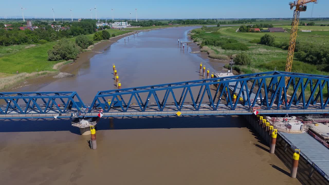Drone shot of the Friesenbrücke railway bridge in closed position over the River Ems. Captures the steel structure, muddy river, and flat countryside on a clear summer day during the building phase.
