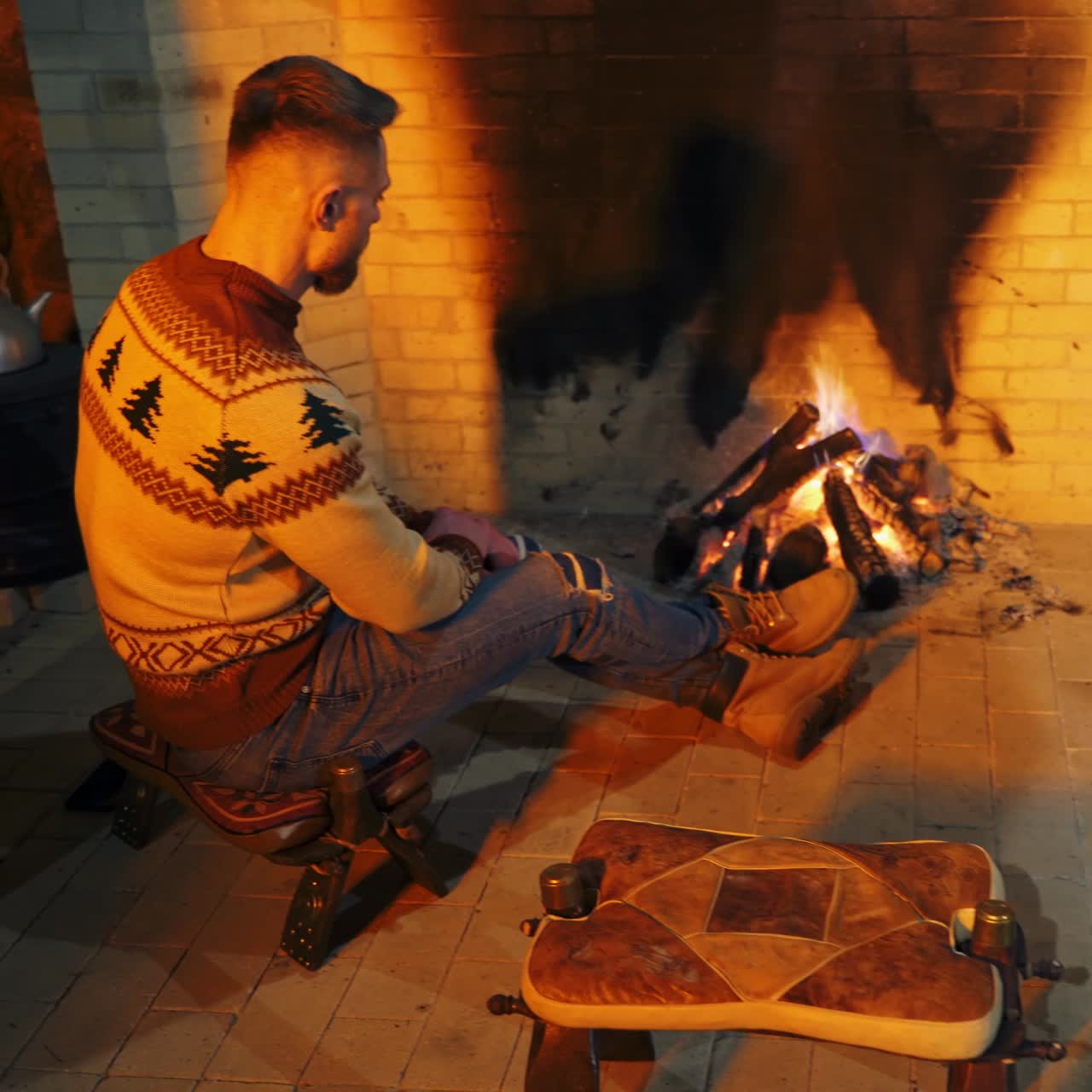 Young bearded guy sit on the chair, rubbing his hands to warm up near fire. Handsome male in sweater inside