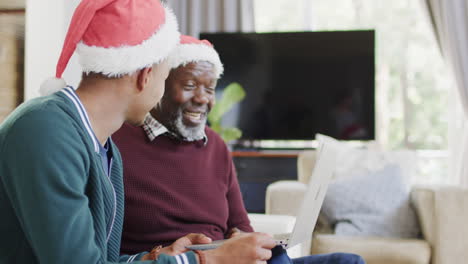 Happy-african-american-father-and-adult-son-in-christmas-hats-having-laptop-video-call,-slow-motion