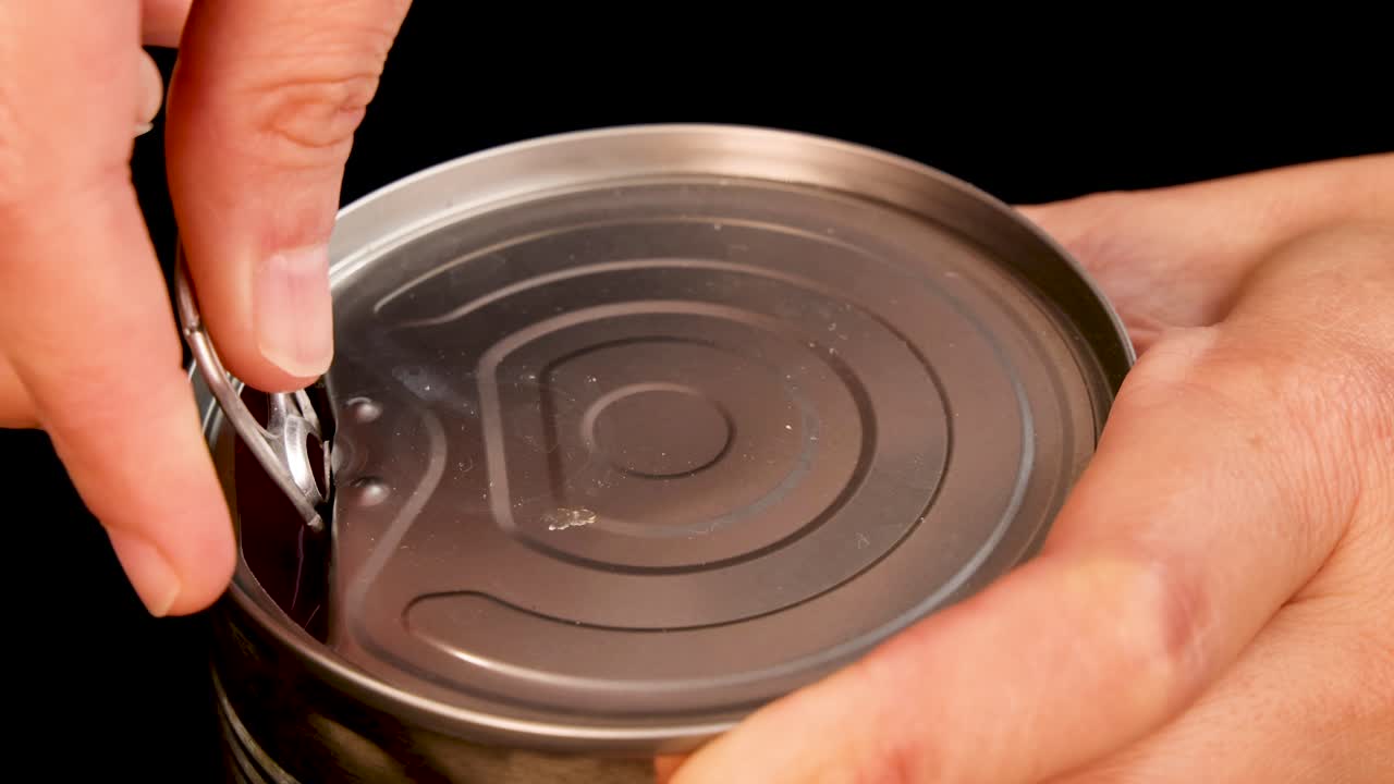 A close-up sequence shows hands opening a can of tomatoes using a pull tab, under bright studio lighting with a black background and steady camera