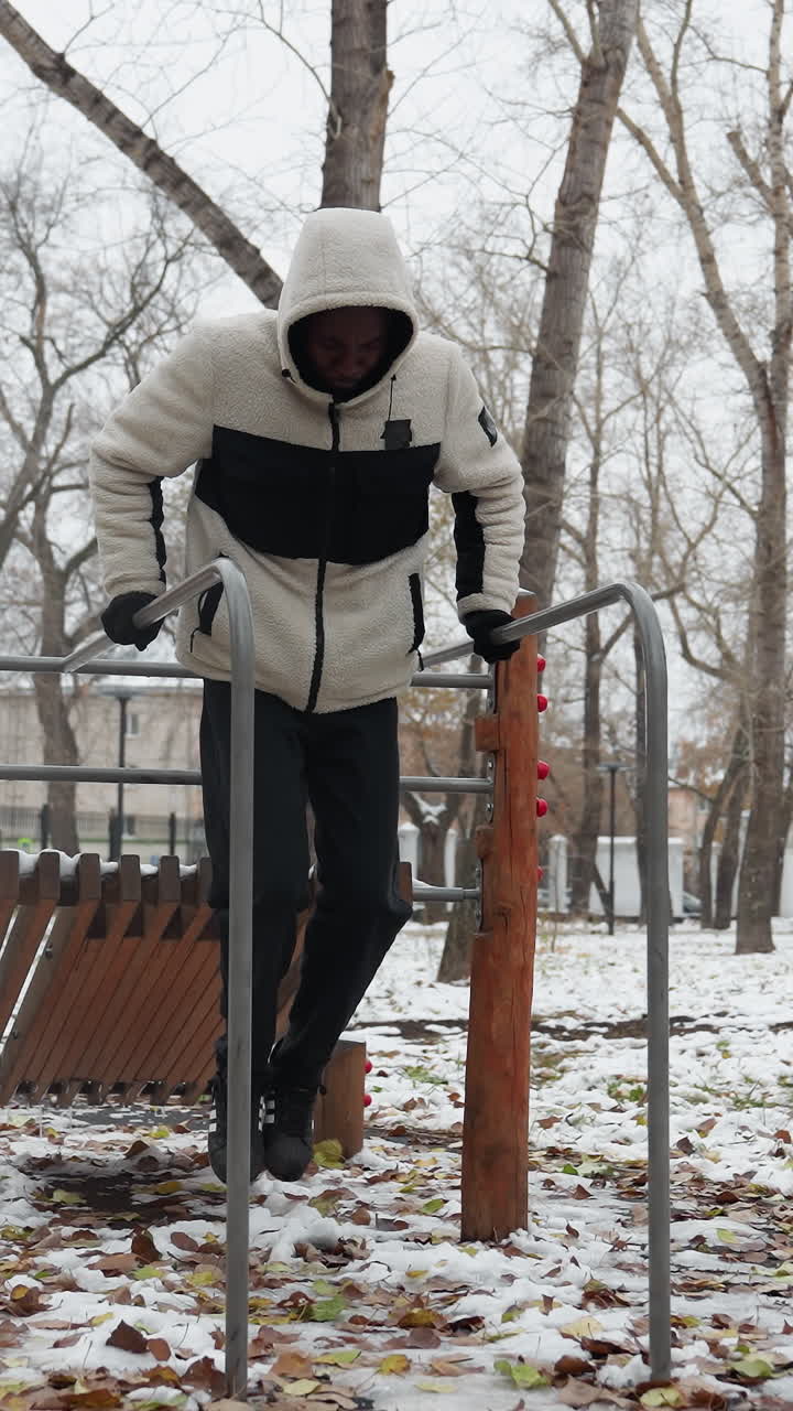 젊은 성인은 눈 인 야외 공원에서 평행 바에서 근육 구축 운동을 수행합니다. 따뜻한 겨울 옷을 입고 차가운 환경에서 운동하는 동안 균형과 힘을 유지합니다.