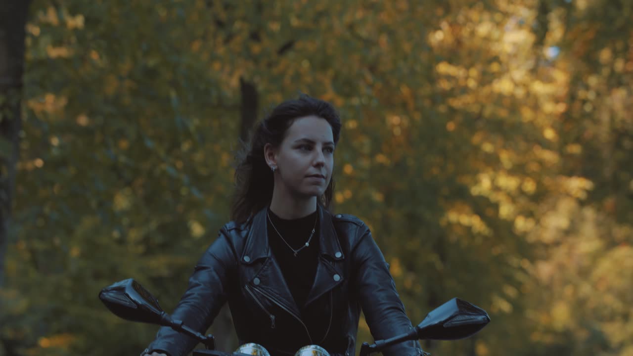una joven europea muy sonriente conduciendo una moto con una chaqueta de cuero en el bosque con hojas de otoño doradas vibrantes y coloridas en un día soleado