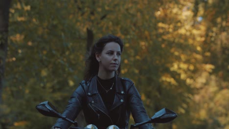 pretty smiling european young woman driving a motorbike wearing leather jacket in forest with vibrant, colorful golden autumn leaves on sunny day