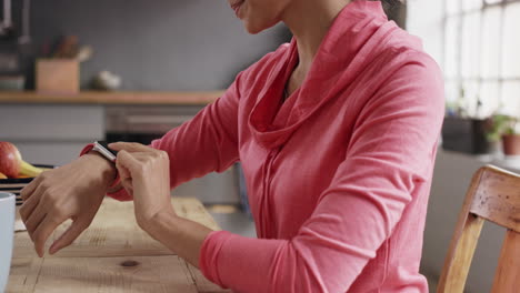 Mixed-race-sporty-woman-using-smart-watch-at-home-drinking-coffee