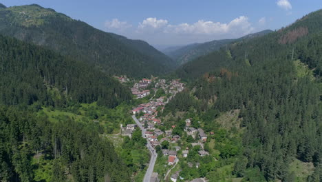 aérea: volando sobre un pequeño pueblo en bulgaria ubicado entre dos colinas, rodeado de bosques de coníferas