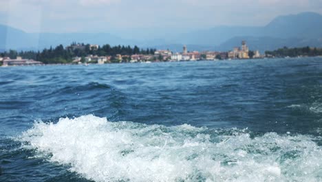 Estela-Y-Lavado-Blanco-Desde-La-Vista-Trasera-Del-Barco-En-El-Lago-De-Garda,-Italia,-Vista-De-La-Costa-Europea-Detrás