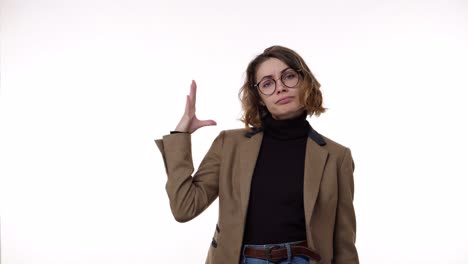 mujer bonita molesta de pie frente a la cámara aislada sobre un fondo blanco y mostrando blah blah gesto, chismes. irritada, emocional. con chaqueta marrón, retro, vaqueros y gafas elegantes