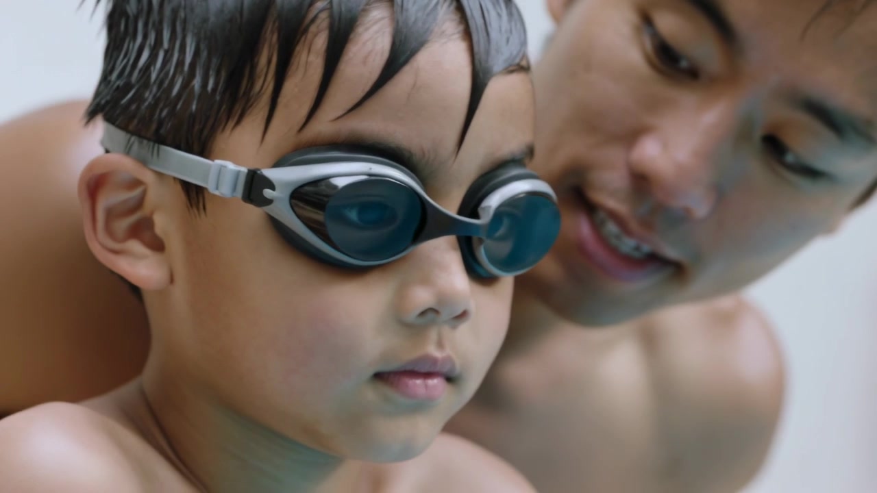 Padre che si china per mettere gli occhialini al bambino in costume a bordo piscina, regolando la cinghia e preparandosi a nuotare.